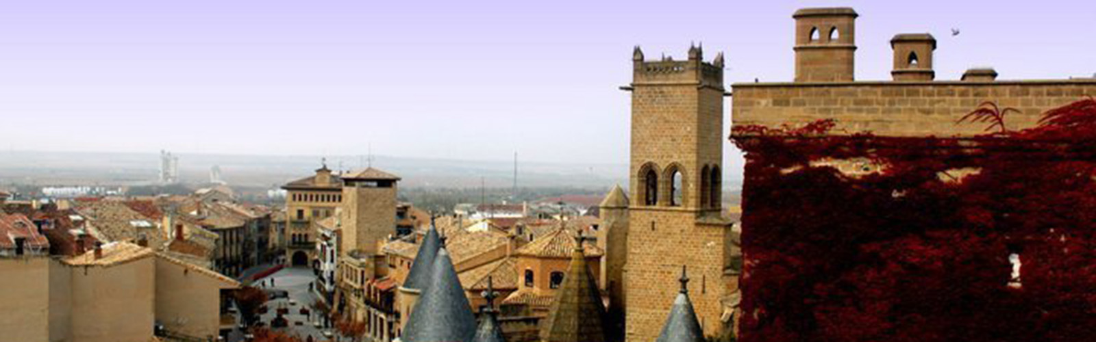 Old buildings of tan bricks line both sides of a road, with a hilly landscape on the horizon.